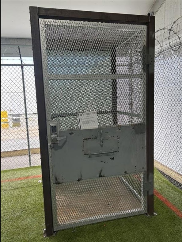 A small metal-mesh enclosure with a solid lower door panel, a food slot and a push-button combination lock, at Alligator Alcatraz.